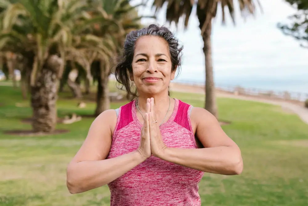 Person in pink athletic top standing outdoors with hands in prayer pose, surrounded by palm trees and greenery