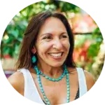 A woman with long brown hair smiles outdoors, wearing a white sleeveless top and turquoise jewelry. The background features lush green foliage and colorful blurred elements, suggesting a garden or tropical setting.