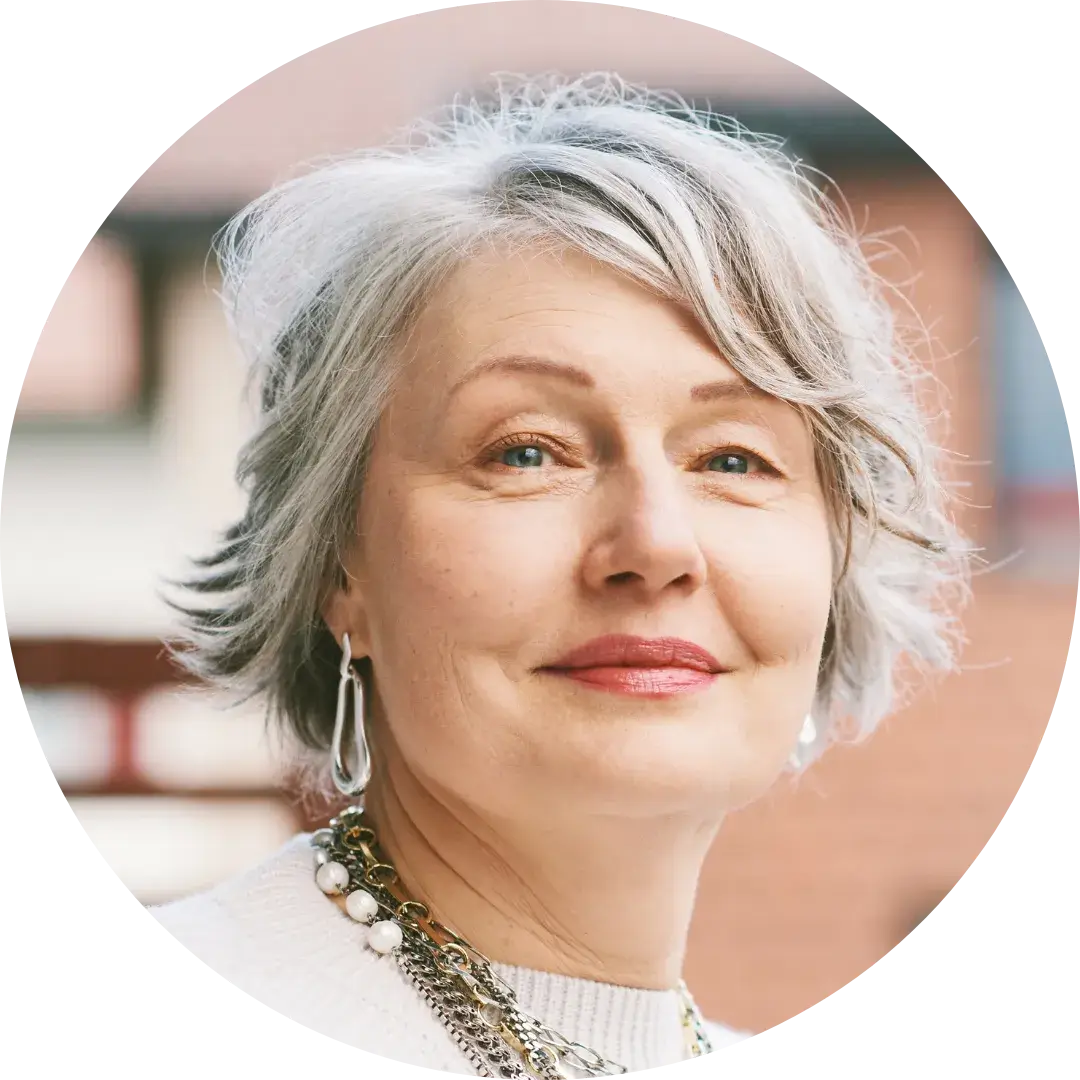 A person with short gray hair wears a white top and a multi-strand necklace. They are looking slightly off-camera with a neutral expression. The background shows an outdoor setting with blurred architectural elements, including red brick and white accents.
