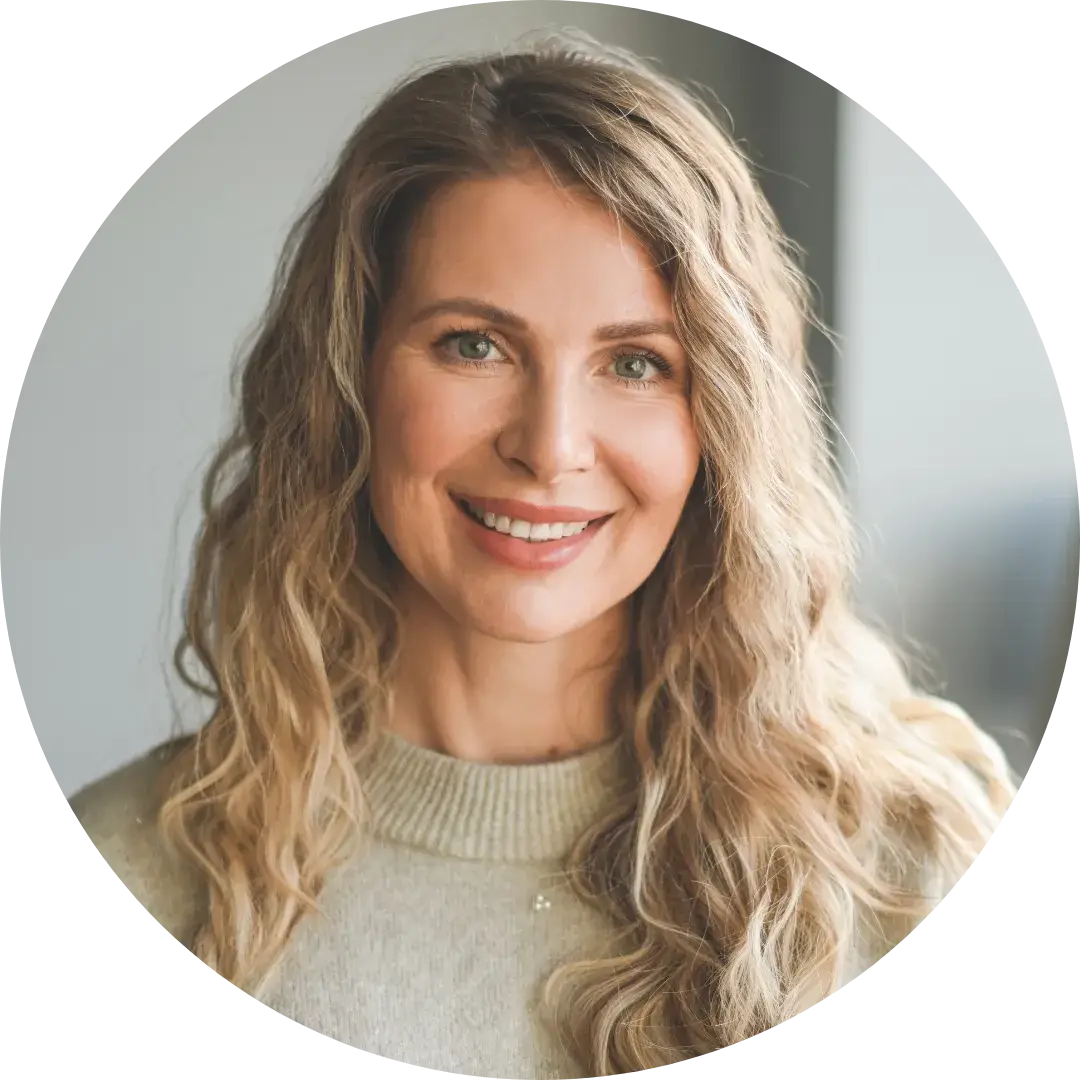 A person with long wavy blonde hair wears a light-colored top and looks directly at the camera with a neutral expression. The softly blurred background suggests an indoor setting with natural light coming through a window.