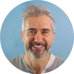 Smiling older adult with gray hair and beard, wearing a gray shirt over a white undershirt, posed against a light blue background.