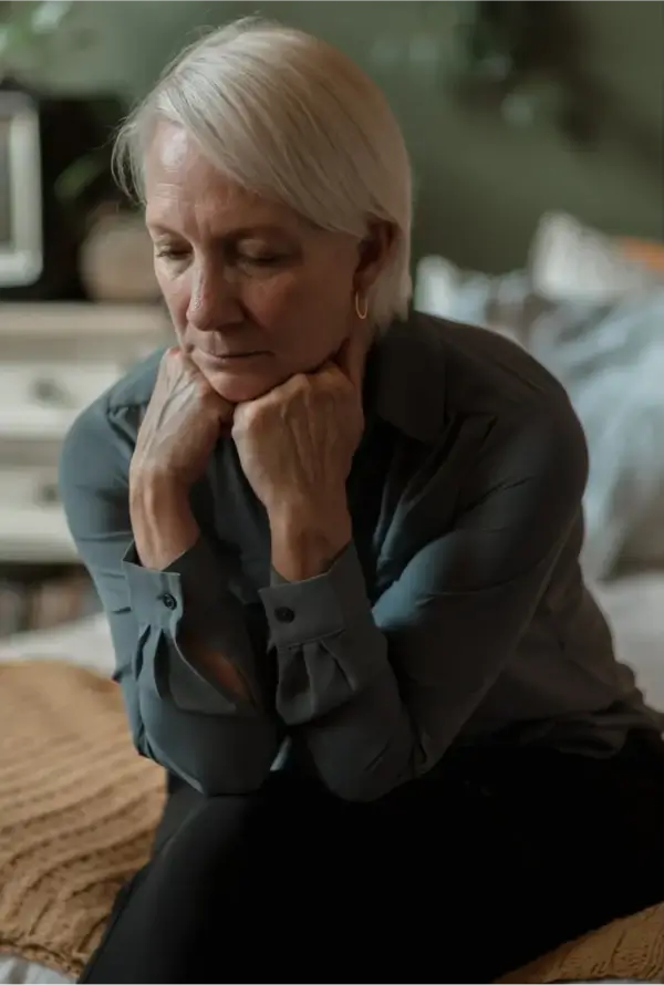 An older adult with short white hair sits on a bed, resting their chin on clasped hands in a contemplative pose. They wear a long-sleeved buttoned shirt and dark pants. The softly lit bedroom setting includes a nightstand, lamp, and decorative items, creating a calm and introspective atmosphere.