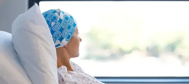 A person wearing a blue and white patterned headscarf and hospital gown sits or lies in a hospital bed, gazing out of a window. Soft lighting and a blurred outdoor view create a calm, reflective atmosphere, suggesting the individual may be undergoing treatment and experiencing a moment of quiet contemplation.
