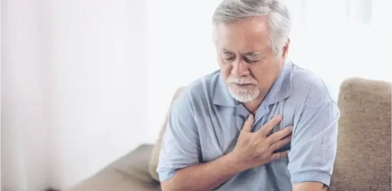 An older adult wearing a light blue polo shirt sits on a couch with a hand placed on their chest, appearing to experience discomfort or pain.