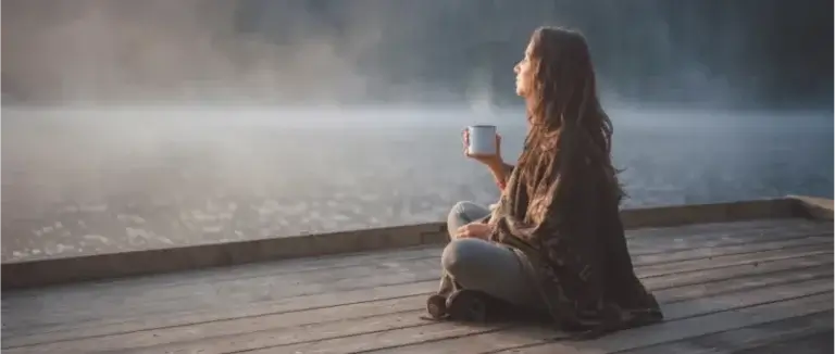 A person sits cross-legged on a wooden dock by a calm, foggy lake, holding a white mug and facing the water. Dressed in casual clothing with a shawl over the shoulders, they appear to be enjoying a peaceful early morning.