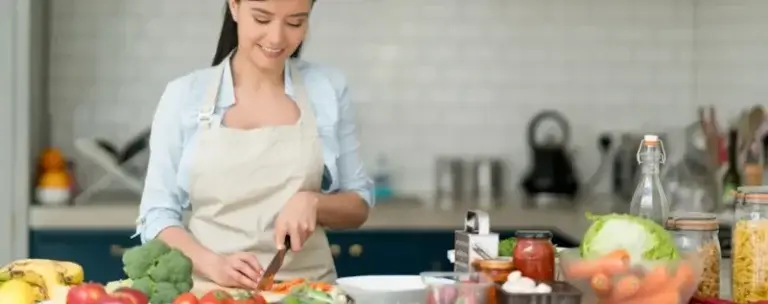 A person wearing a light blue shirt and beige apron slices vegetables on a cutting board in a kitchen. The counter is filled with fresh produce including broccoli, carrots, tomatoes, bananas, lettuce, and mushrooms, along with jars of pasta, a bottle of oil, and cooking utensils. The background features a tiled backsplash, a kettle, and other kitchen tools, creating a vibrant, health-focused cooking environment.