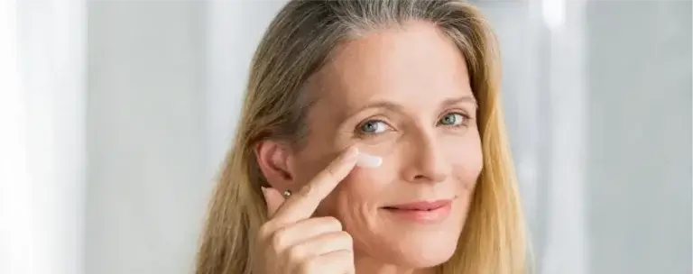 A person with long blonde hair applies a small amount of cream under the right eye using the index finger. They smile gently and look directly at the camera. The softly blurred background features light-colored curtains, creating a clean and serene atmosphere. The image illustrates a skincare routine, specifically the application of eye cream.