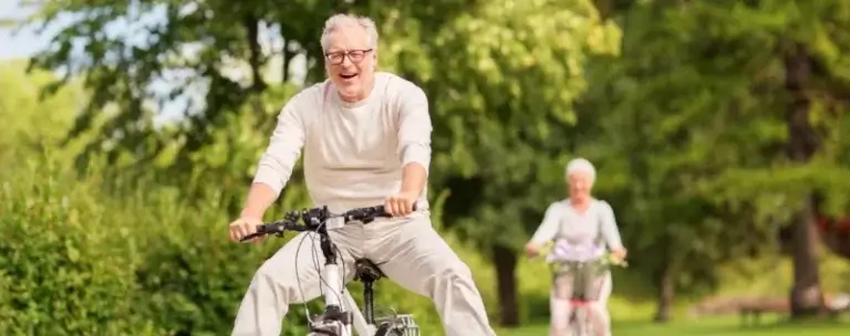 Two older adults ride bicycles along a paved path surrounded by lush greenery. The person in the foreground wears a light-colored outfit and glasses, smiling while cycling. Another cyclist, also dressed in light colors, follows behind. The setting appears to be a park or natural area, highlighting outdoor recreation and active aging.