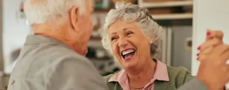 Two older adults share a joyful moment indoors, possibly dancing together. The woman, smiling broadly, holds the man's hand and faces the camera. She has gray curly hair and wears a green cardigan over a pink collared shirt. The man, seen from the side and back, has short white hair and wears a gray shirt. The background includes shelves and cabinets, suggesting a home setting.