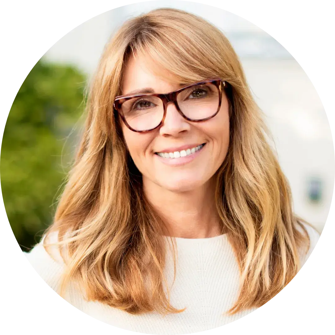 A person with long light brown hair and glasses smiles while wearing a white top. The background is softly blurred, showing greenery and a light-colored building.