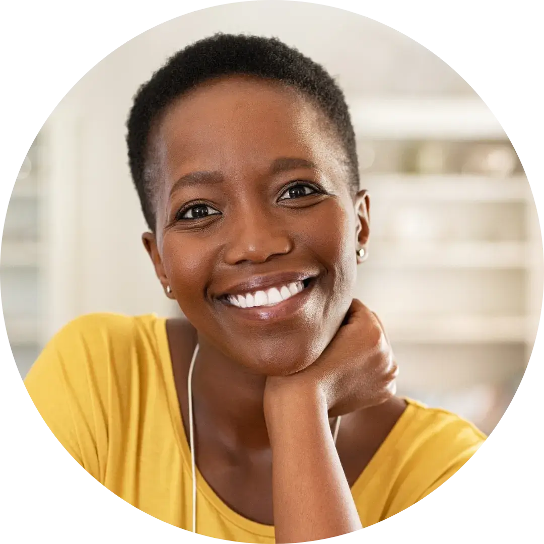A person with short hair smiles while resting their head on one hand. They are wearing a yellow top, and the softly focused indoor background includes shelves and household items, suggesting a cozy living space.