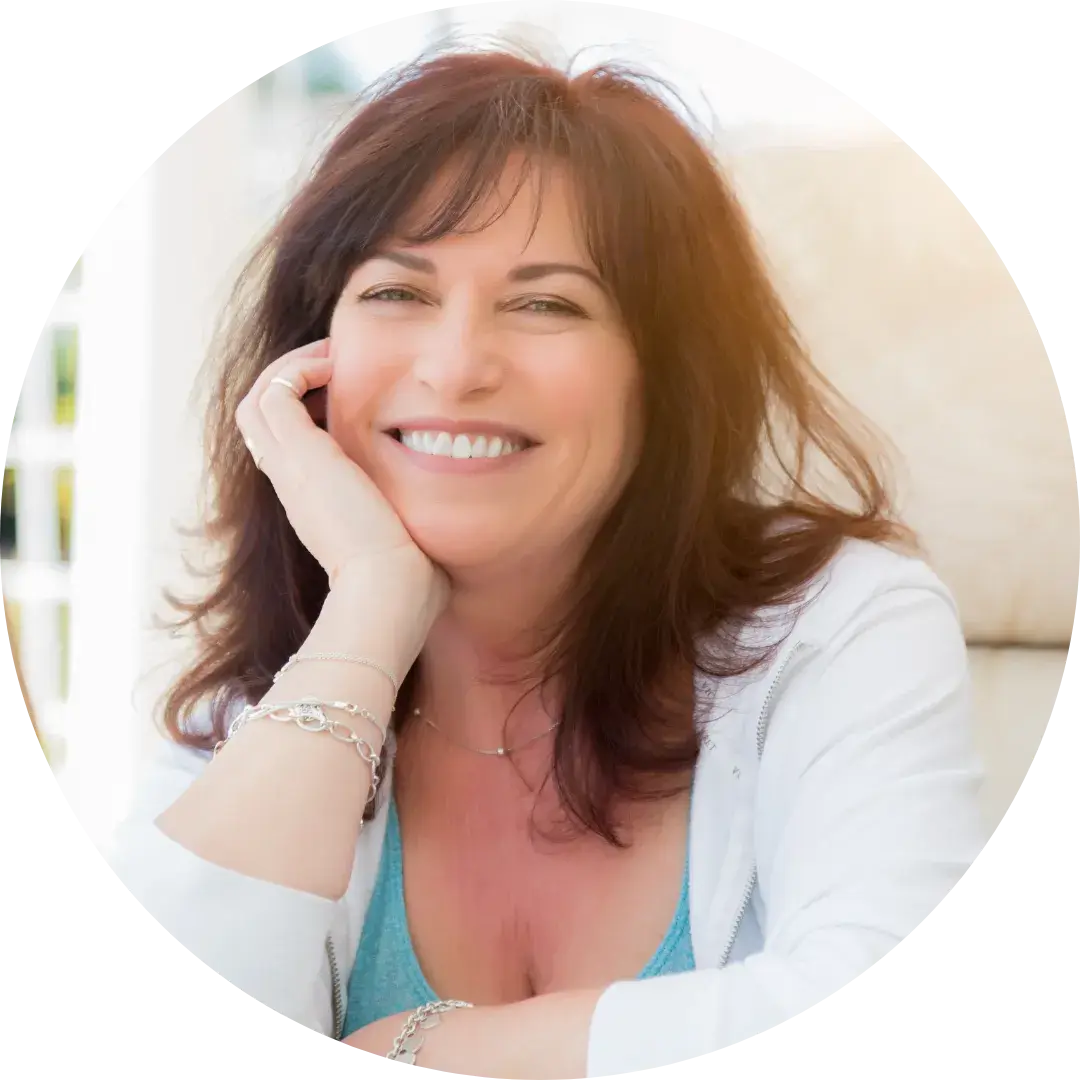A person with long dark brown hair smiles while resting their head on one hand. They are wearing a white top layered over a light blue shirt. The background shows a bright indoor setting with soft furnishings.