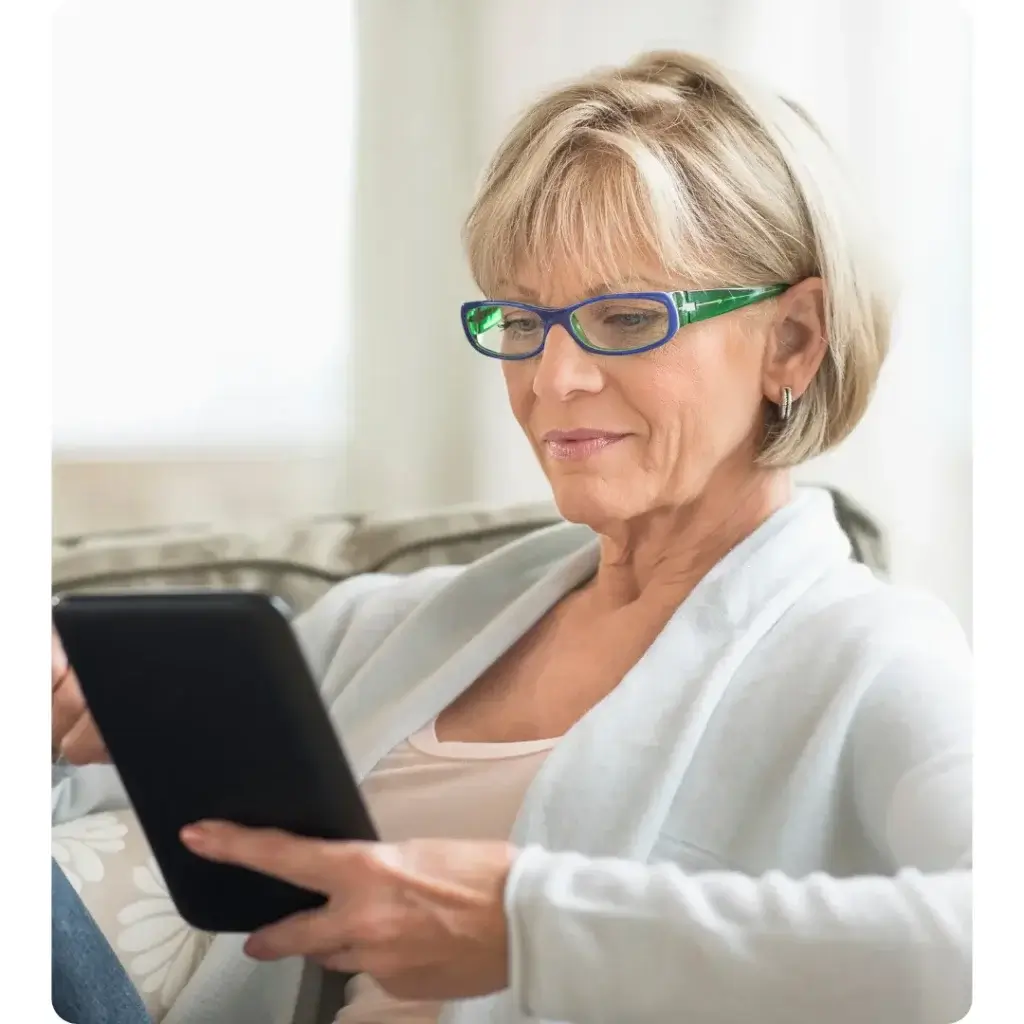 Older adult wearing green and blue eyeglasses sits on a couch, looking at a tablet. They wear a light cardigan over a pink top. A floral pillow and light curtain are visible in the background.