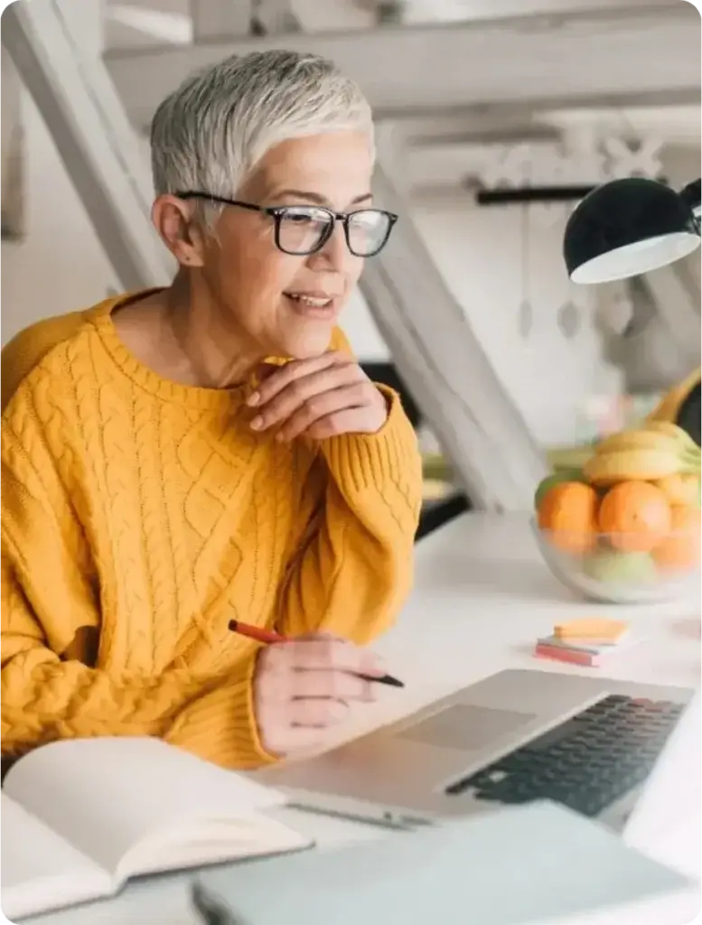 Older woman with short gray hair and glasses taking notes while she watches something on her laptop