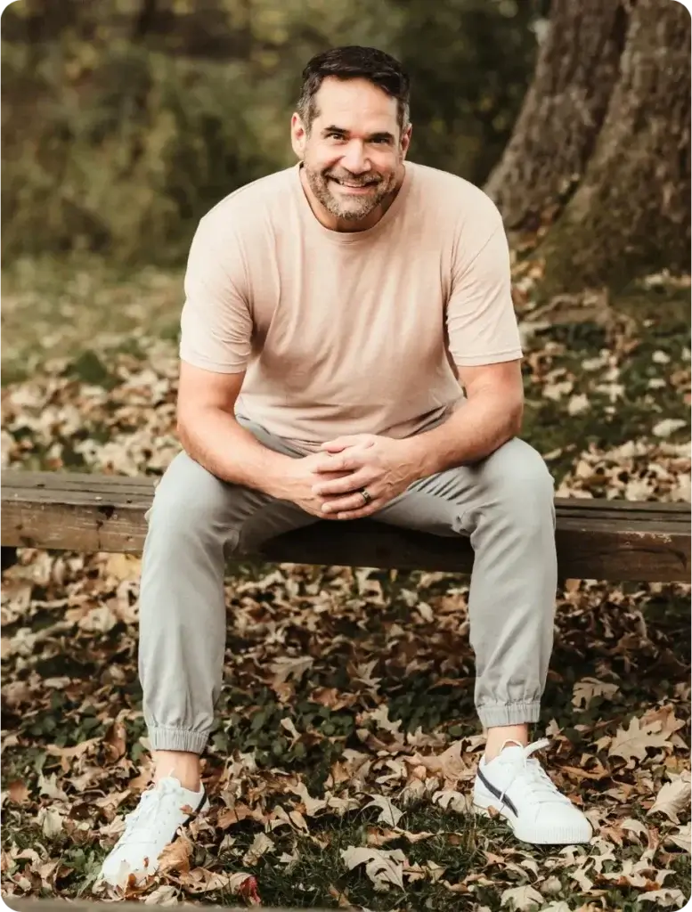 Smiling man in a tan T-shirt sitting on a wooden bench outdoors, surrounded by fallen autumn leaves